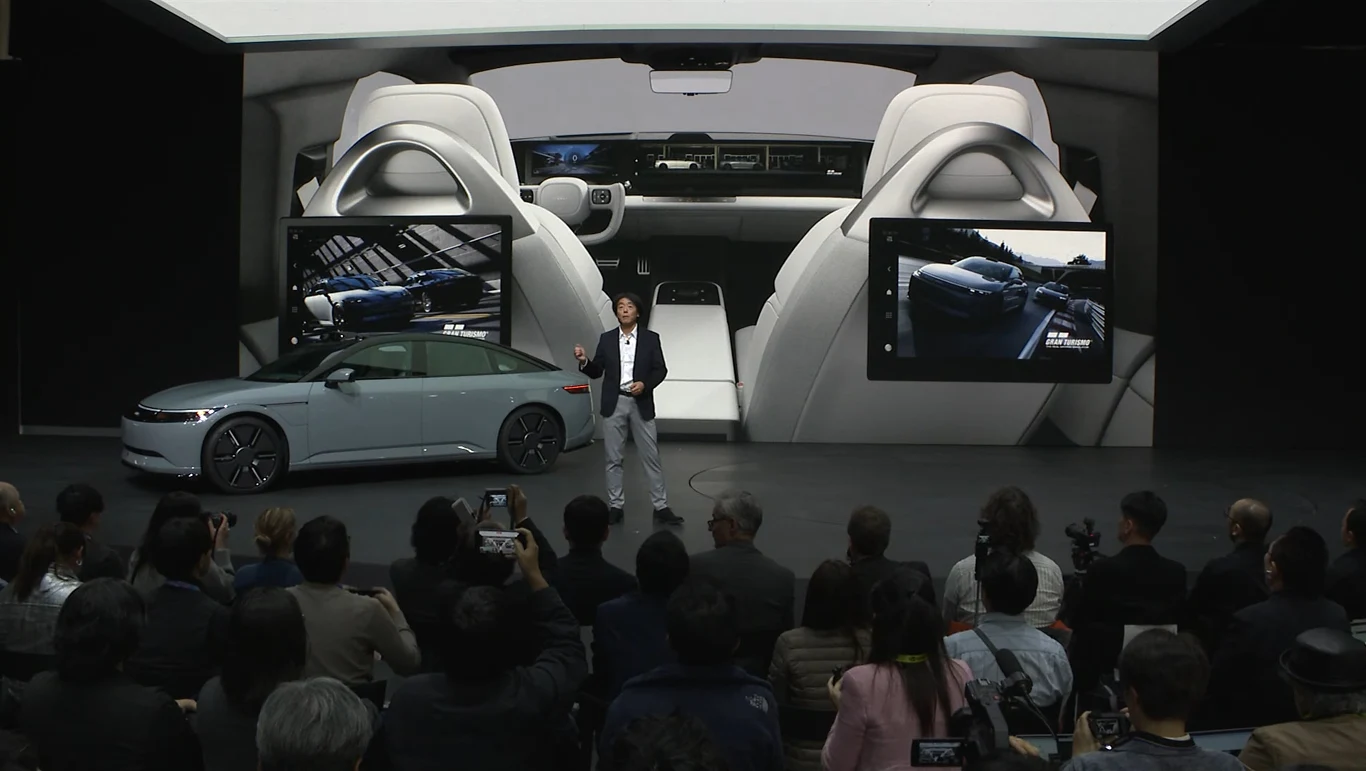 Poza modyfikacją sfery wizualnej Afeela 1 zaoferuje także opcje personalizacji audio. Ciekawe, czy będzie można wybierać dźwięk silnika z pomiędzy motoryzacyjnych legend dostępnych w serii Gran Turismo Konferencja prasowa poświęcona premierze nowego modelu samochodu elektrycznego, przemawiający mężczyzna stoi przed samochodem na tle dużego ekranu z wizualizacją wnętrza pojazdu, widownia dokumentuje wydarzenie smartfonami.