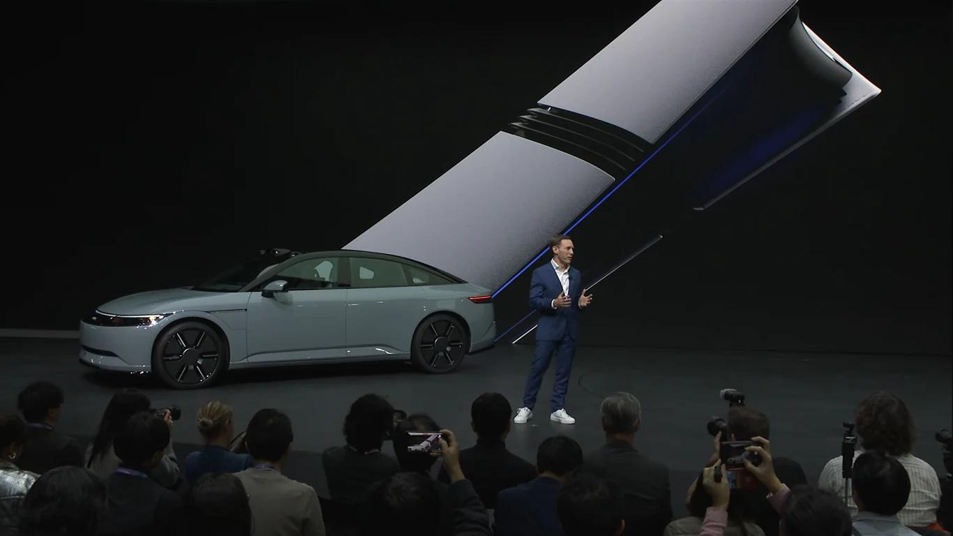 Afeela 1 prawie jak PlayStation 5 na kołach Mężczyzna w garniturze prezentuje nowoczesny samochód elektryczny na tle dużego ekranu z grafiką elektrycznego przewodu, wokół zgromadzona publiczność, wiele osób robi zdjęcia.