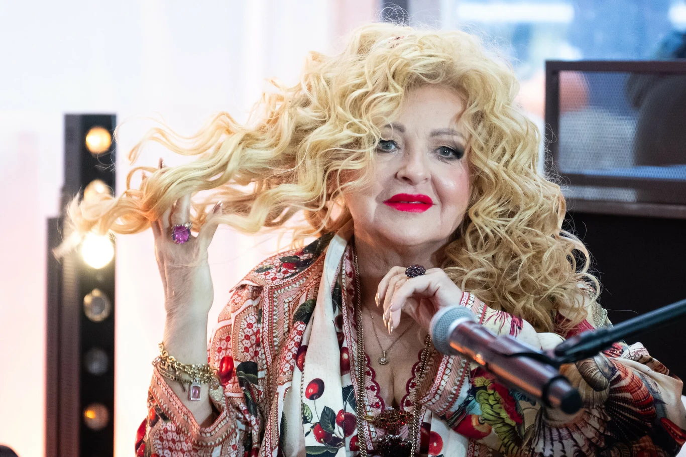 Kobieta o blond, kręconych włosach w kolorowym stroju siedzi przy mikrofonie, delikatnie dotykając swoich włosów, z wyrazistym makijażem, na tle elementów oświetleniowych.