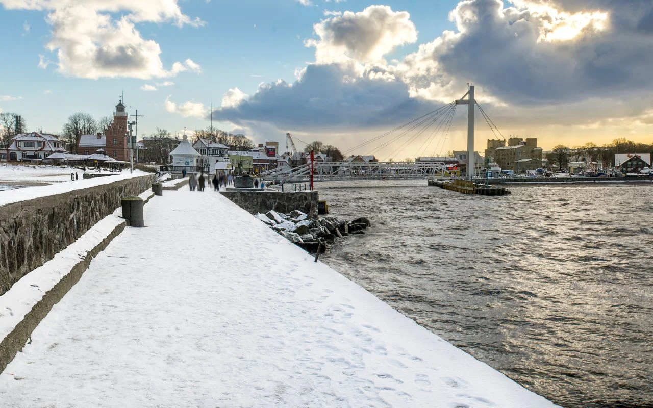 Zaśnieżony deptak wzdłuż rzeki prowadzący w stronę charakterystycznego mostu wiszącego. Po lewej stronie widoczne są budynki oraz kościół z czerwoną wieżą. Nad wodą unosi się dynamiczne, zimowe niebo z promieniami słońca przebijającymi się przez chmury.