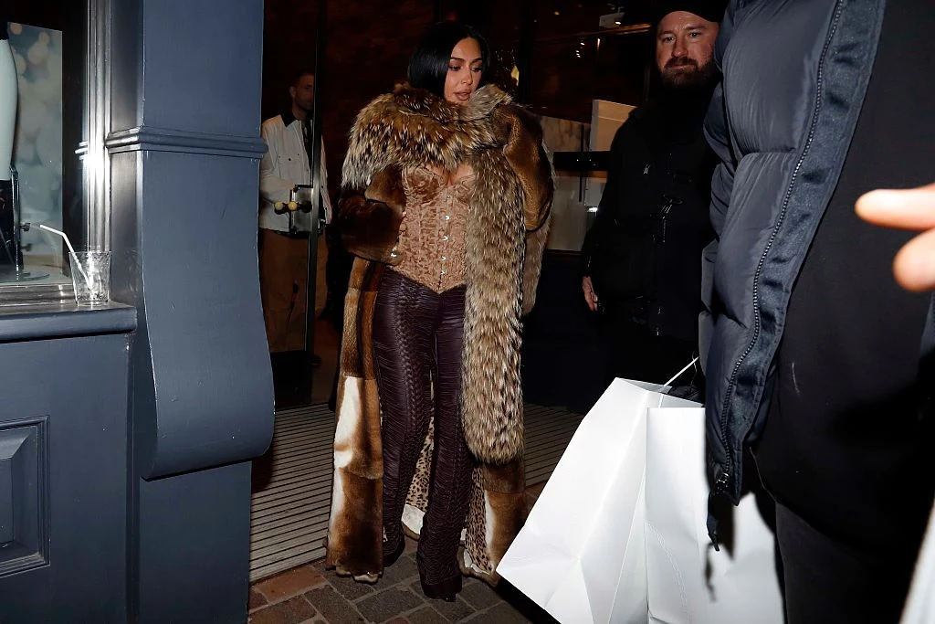 Kobieta ubrana w efektowne futro oraz błyszczącą gorsetową bluzkę stoi na tle wejścia do lokalu, obok niej znajdują się osoby towarzyszące, jedna osoba trzyma duże białe torby zakupowe.