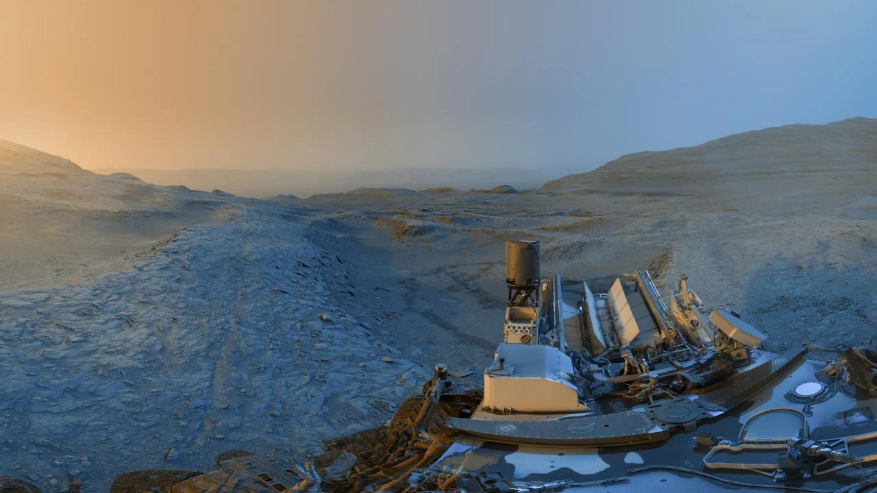 Panorama Marsa uchwycona przez łazik Curiosity agencji NASA w listopadzie. Marsjański krajobraz z widocznym łazikiem NASA na pierwszym planie, w tle pagórkowaty, pustynny teren oraz zmieniające się światło nieba od pomarańczowego do niebieskiego.