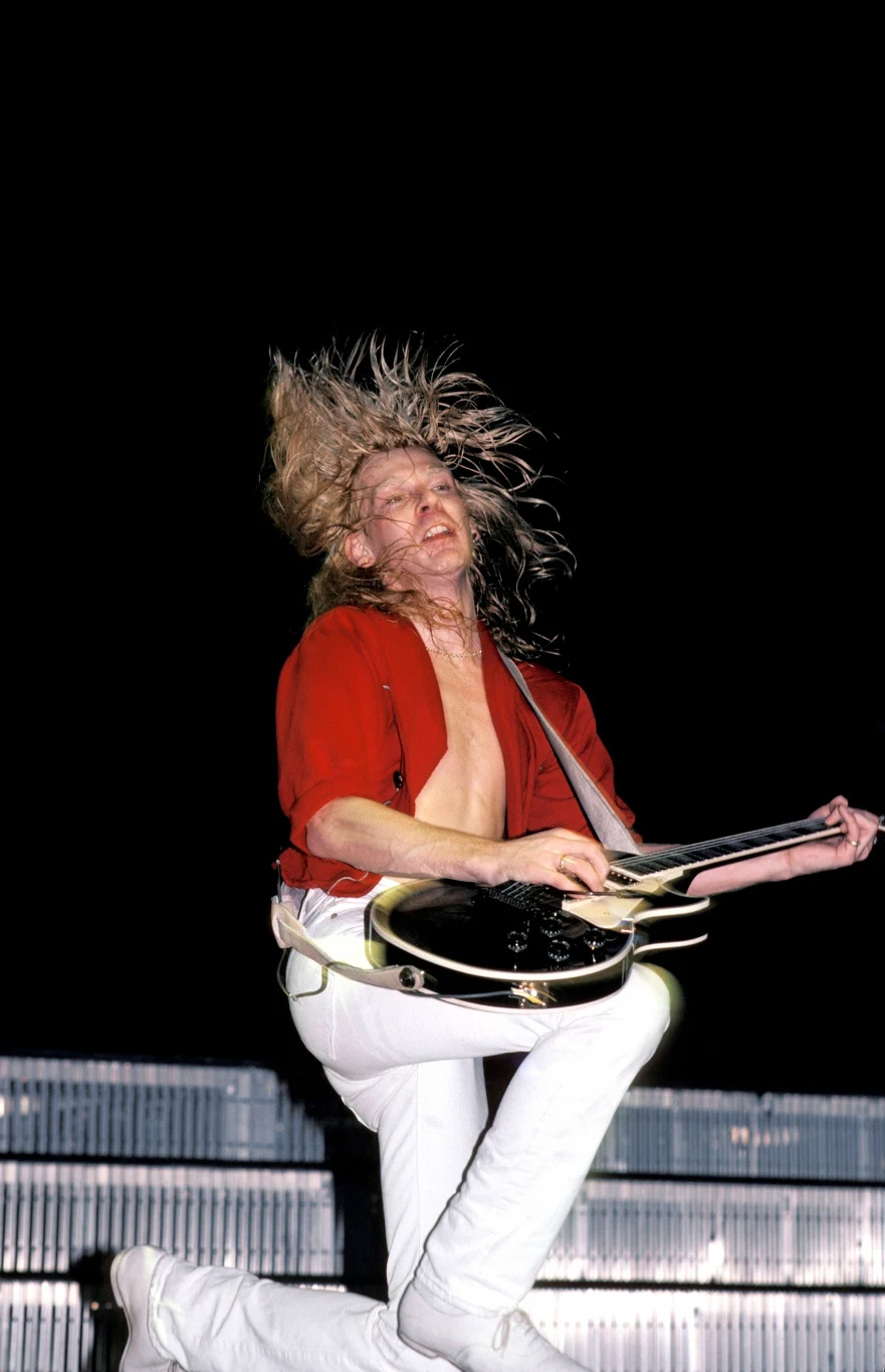 Mężczyzna z długimi, blond włosami ubrany w czerwoną koszulę i białe spodnie, skacze w powietrzu z gitarą elektryczną podczas koncertu, dynamiczna poza i intensywny wyraz twarzy podkreślają energię występu, tło ciemne, scena oświetlona światłem scenicz...