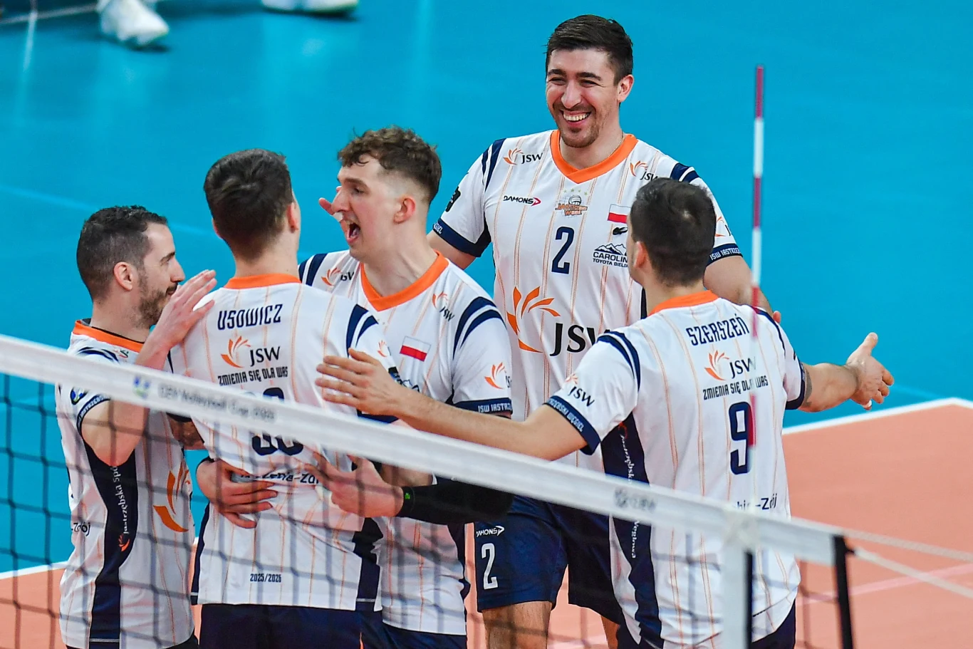 Siatkarze JSW Jastrzębskiego Węgla z awansem w Pucharze CEV Grupa siatkarzy w białych koszulkach z niebieskimi elementami celebruje punkt na boisku, uśmiechnięci zawodnicy obejmują się i okazują radość w trakcie meczu siatkarskiego.