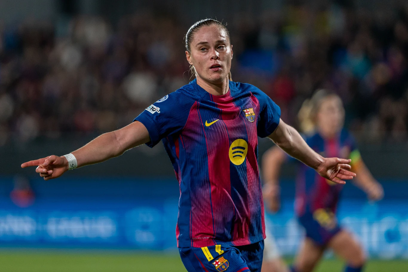 Zawodniczka piłki nożnej w stroju FC Barcelona wykonuje gest wskazujący podczas meczu, w tle widoczne zamazane sylwetki innych zawodniczek i trybuny.
