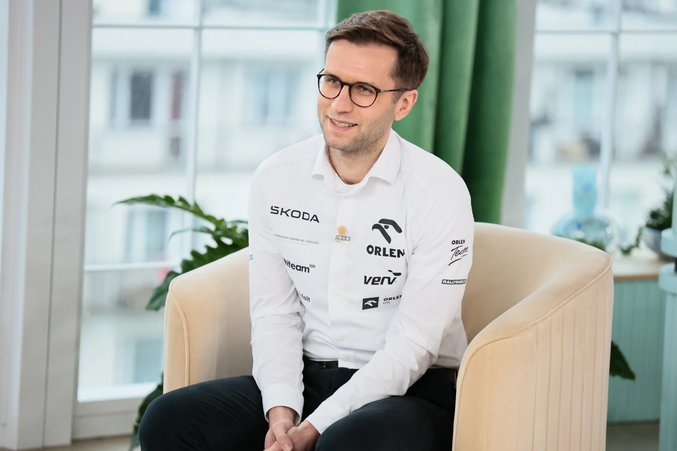 Mężczyzna w okularach siedzi na jasnym, wygodnym fotelu, ubrany w białą koszulę z logotypami sponsorów sportowych. W tle duże okno z widokiem na zabudowania, zielona roślina oraz jasne zasłony.