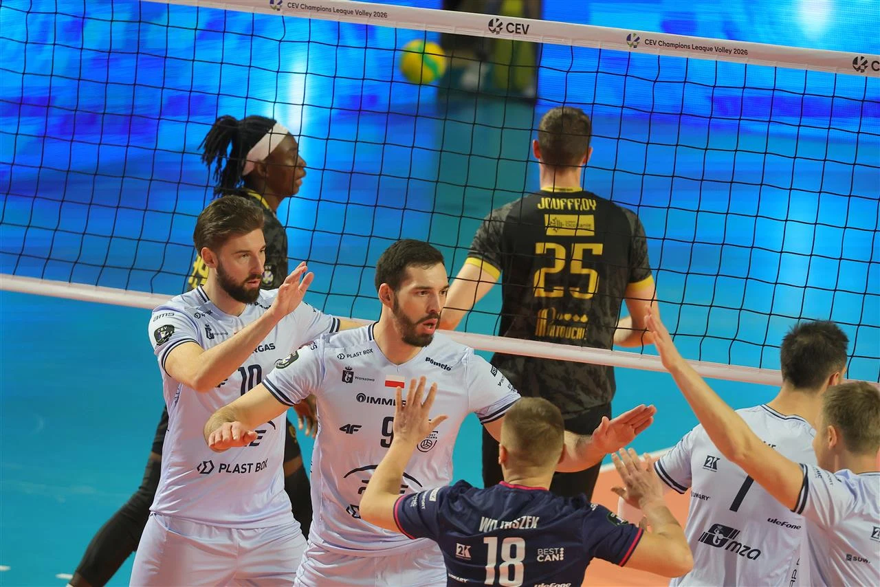 Zawodnicy drużyny siatkarskiej w białych strojach wykonują gest przybijania piątek po udanej akcji na boisku. Po drugiej stronie siatki widoczni rywale w czarnych koszulkach, dynamiczne oświetlenie podkreśla sportową atmosferę.