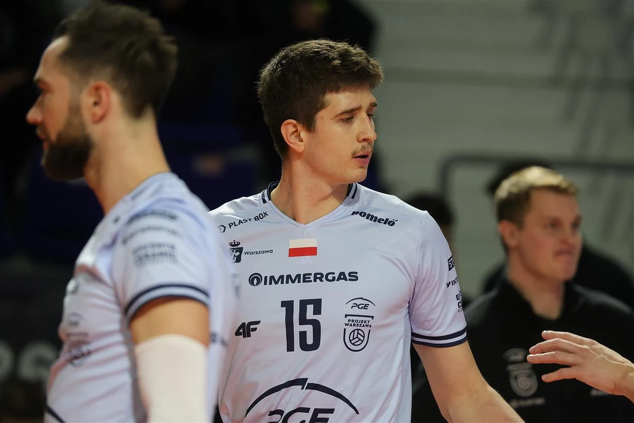 Siatkarz ubrany w jasny sportowy strój z numerem 15 oraz polską flagą na piersi stoi na boisku, obok widoczny jest inny zawodnik oraz sylwetka osoby w tle.