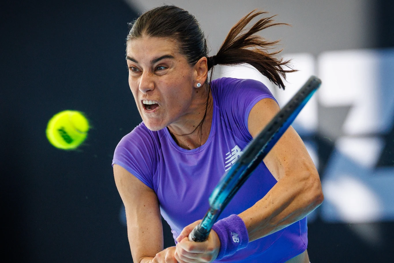 Tenisistka uderzająca piłkę rakietą podczas intensywnego meczu, z wyrazem skupienia i determinacji na twarzy, w fioletowej koszulce i opasce na nadgarstku, z dynamicznym ruchem włosów i unoszącą się piłką.