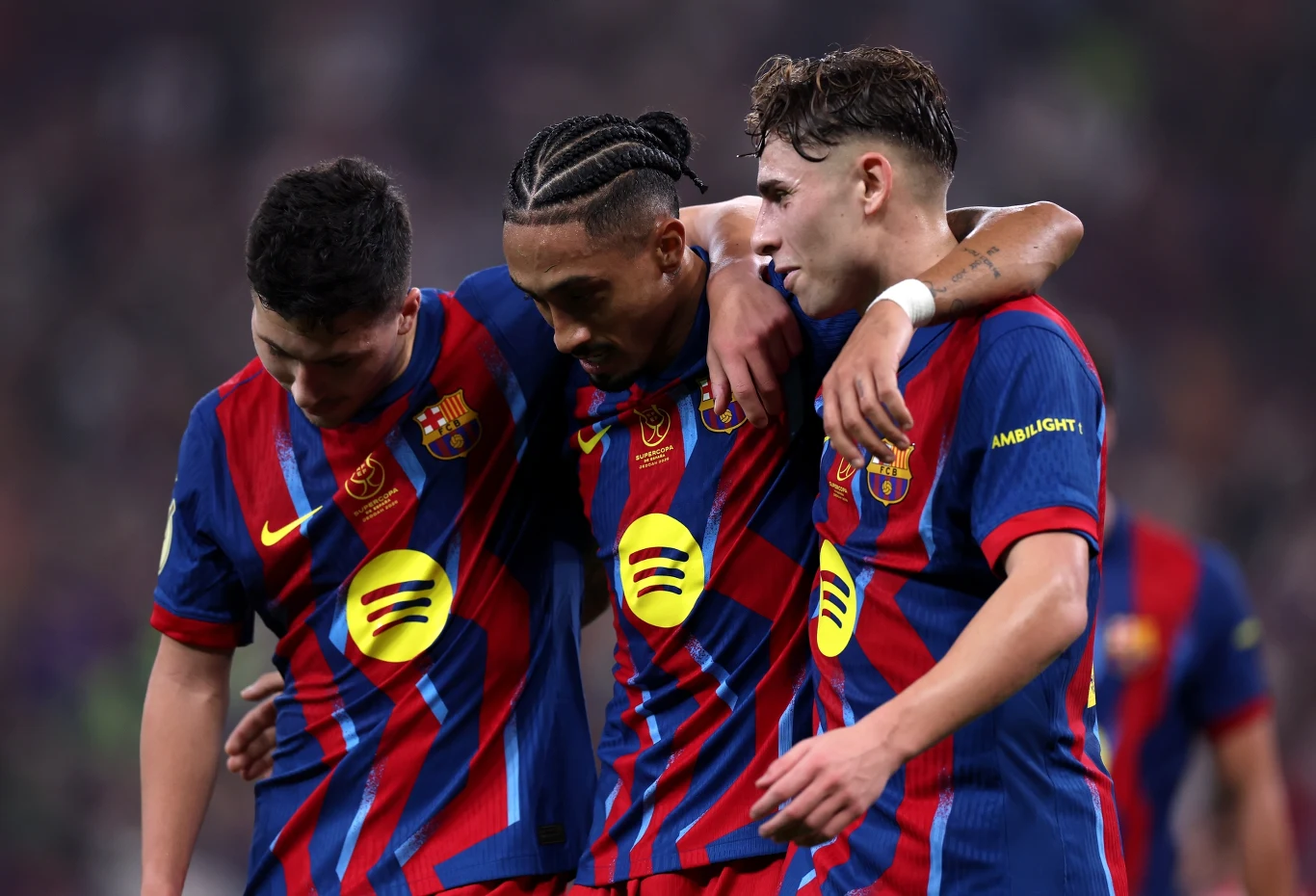 FC Barcelona w meczu z Athletikiem Trzech piłkarzy w koszulkach FC Barcelony obejmuje się ramionami, wykazując wsparcie i koleżeństwo na boisku. Młodzi mężczyźni noszą charakterystyczne niebiesko-granatowe stroje z żółtymi detalami oraz herbem klubu.