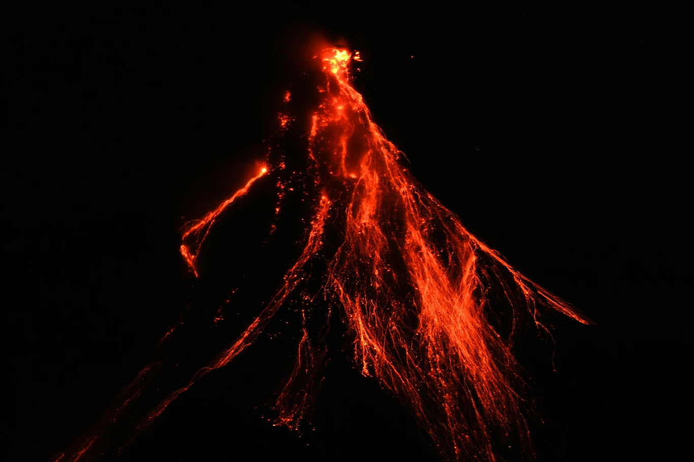 Erupcja wulkanu z wyrzucającą jasnoczerwoną lawą, rozświetlającą ciemność nocnego nieba, z wyraźnie widocznymi strumieniami gorącej materii spływającymi po zboczach wulkanu.