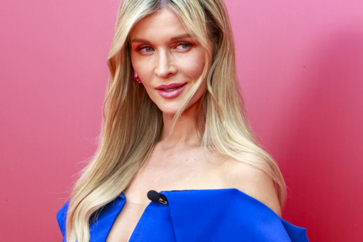 Joanna Krupa Kobieta o długich blond włosach, ubrana w elegancką suknię w kolorze intensywnego niebieskiego, pozuje na tle różowej ściany. Uwagę przyciąga jej delikatny makijaż oraz subtelny uśmiech.