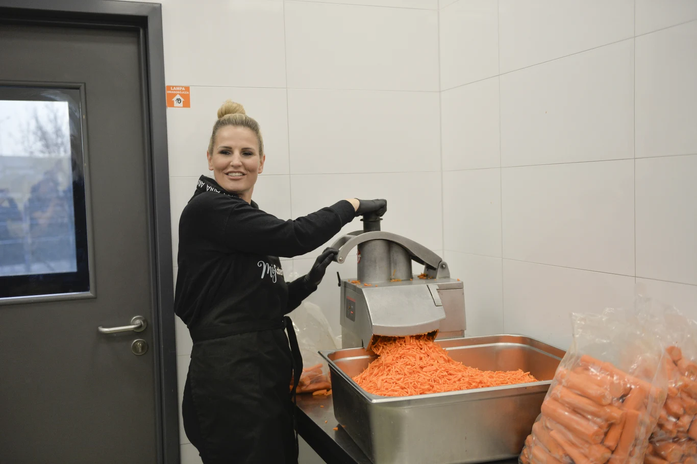 Dominika Tajner, 2022 rok Kobieta ubrana w fartuch obsługuje maszynę do rozdrabniania marchewek, wsypując starte warzywa do dużego stalowego pojemnika; obok znajdują się worki z marchewkami w całości, a tło stanowi jasna kafelkowana ściana.