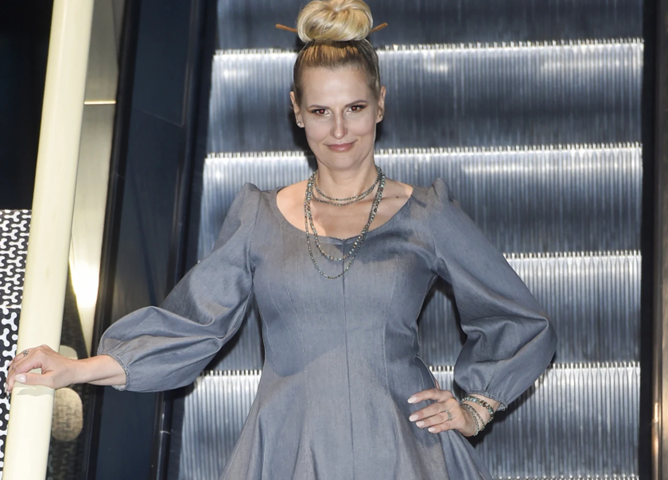 Dominika Tajner, 2021 rok Kobieta w eleganckiej, szarej sukience z balonowymi rękawami stoi na tle ruchomych schodów, z włosami upiętymi w kok oraz licznymi naszyjnikami i bransoletkami, prezentuje się w pewnej pozycji.
