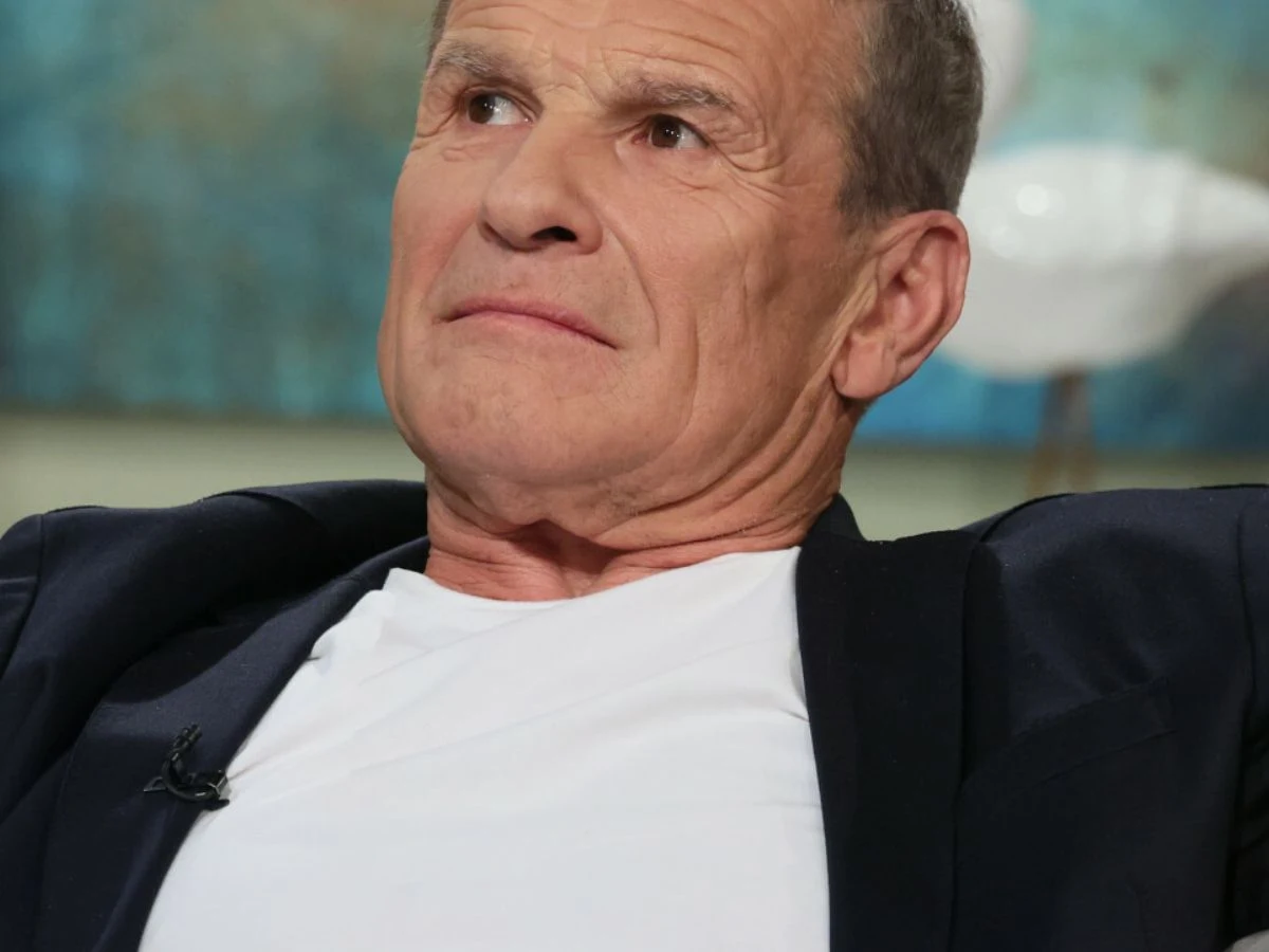 Marek Włodarczyk Mężczyzna w średnim wieku ubrany w biały t-shirt i ciemną marynarkę, siedzący w studiu telewizyjnym na tle rozmazanego obrazu w niebieskich barwach, ze spokojnym wyrazem twarzy.