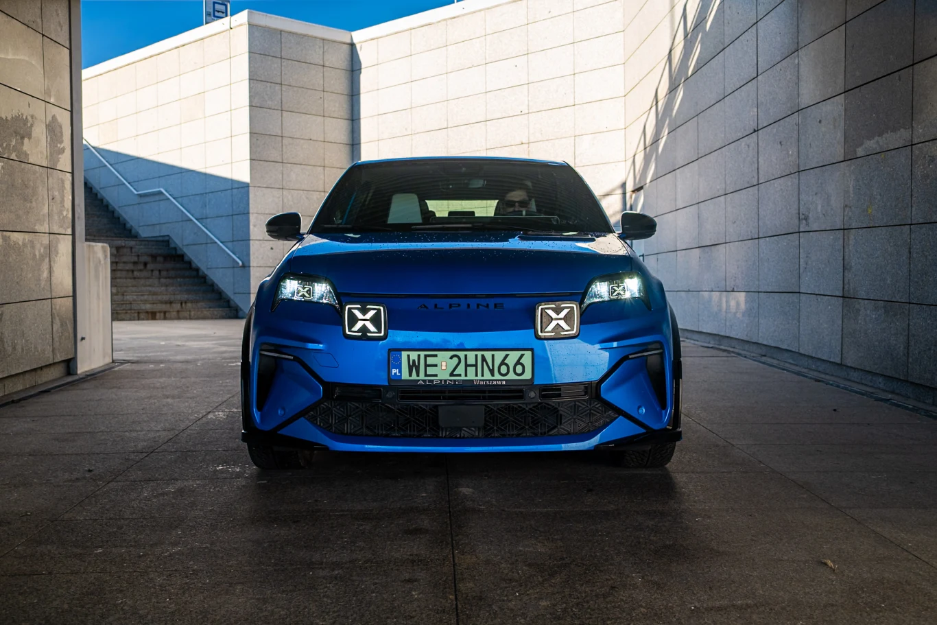 Niebieski samochód sportowy ustawiony centralnie, widoczny od frontu, na tle betonowych ścian i schodów. Auto ma wyraziste światła oraz polskie tablice rejestracyjne. Sylwetka pojazdu podkreślona ostrymi liniami nadwozia.