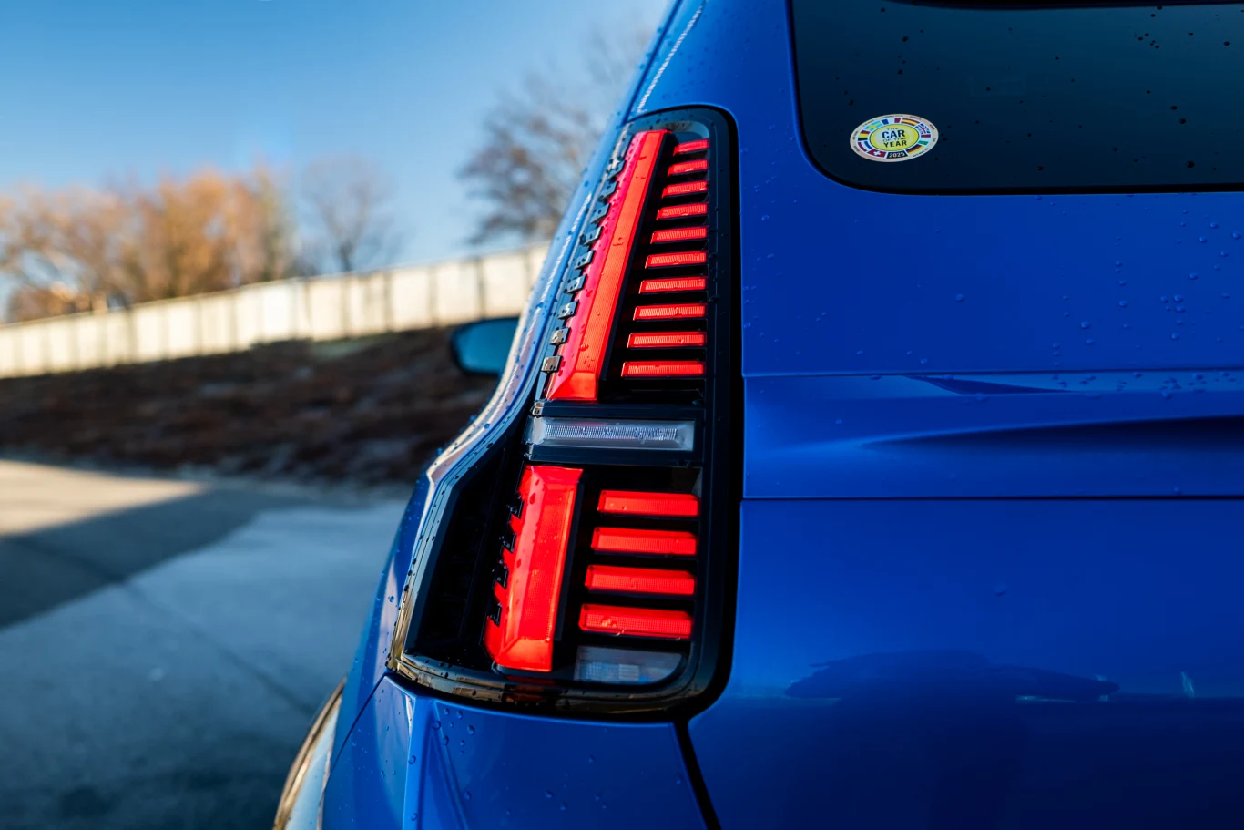 Tylna lampa LED niebieskiego samochodu osobowego z charakterystycznymi czerwonymi światłami, na szybie widoczna okrągła naklejka; w tle rozmazany płot i drzewa.
