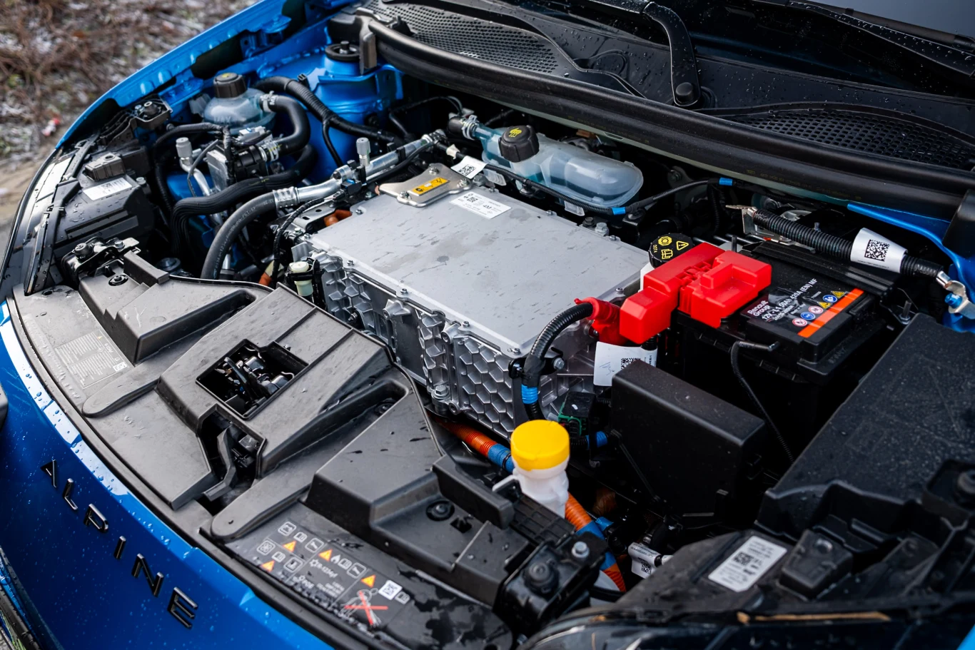 Komora silnika samochodu elektrycznego z widocznymi podzespołami, przewodami, akumulatorem i oznaczeniami bezpieczeństwa, dominujący srebrny moduł elektryczny oraz charakterystyczne niebieskie elementy eksploatacyjne.