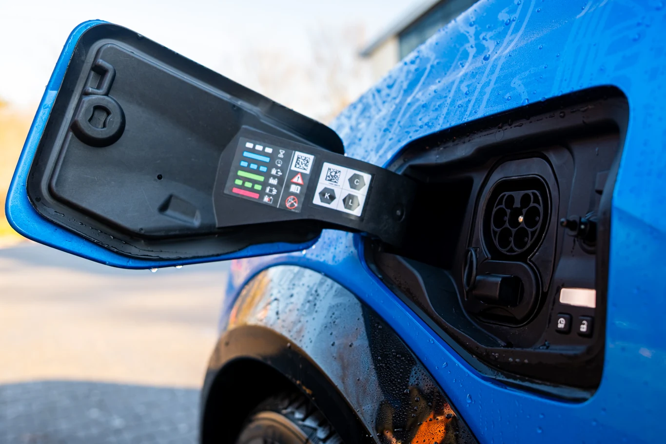 Wlew do ładowania samochodu elektrycznego w niebieskim pojeździe, z wyraźnie widocznym gniazdem oraz naklejkami informacyjnymi na otwartej klapce wlewu, na powierzchni auta widoczne krople wody.