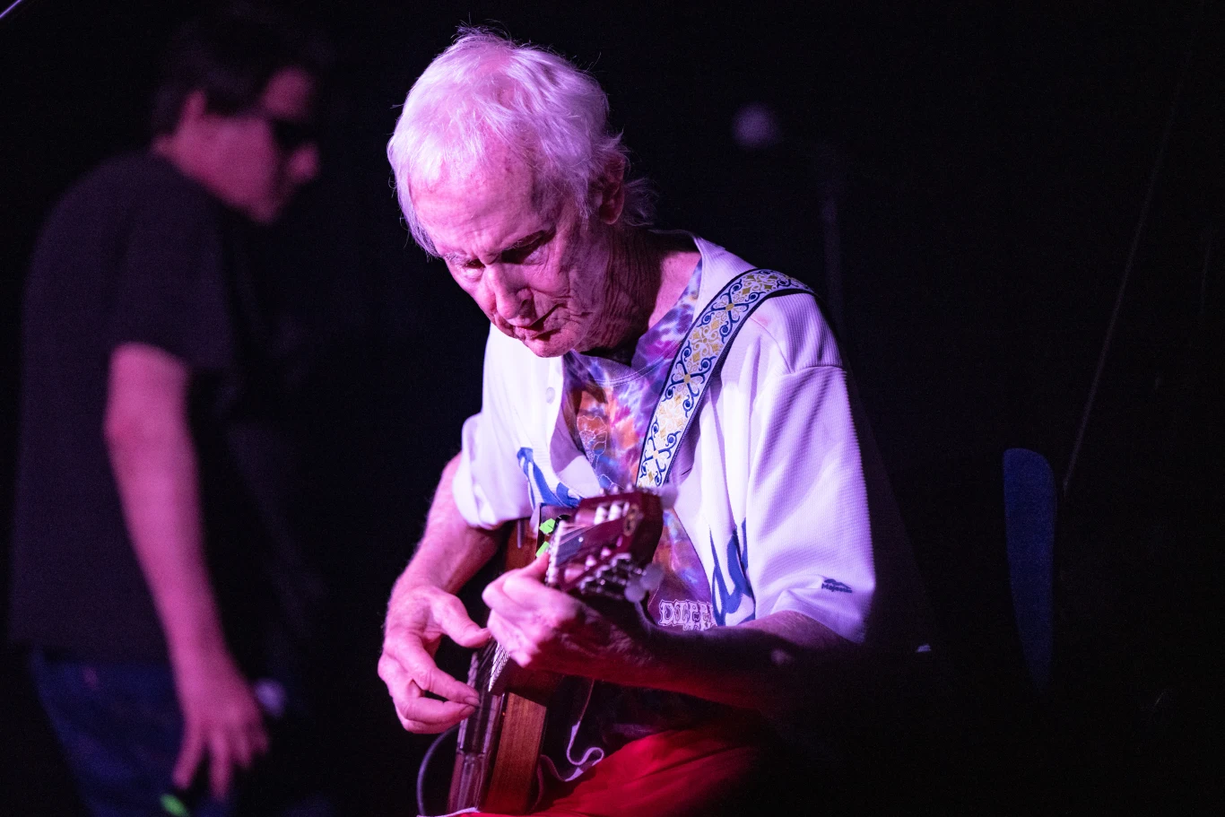 Starszy mężczyzna gra na gitarze elektrycznej na scenie, skupiony na muzyce, z widocznym kolorowym paskiem od gitary, w tle druga osoba lekko zamazana, scena oświetlona kolorowym światłem.