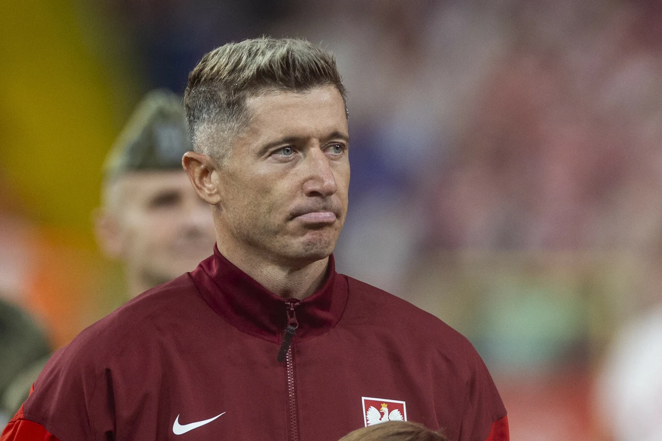 Mężczyzna w bordowej kurtce z godłem Polski na piersi, stoi na tle rozmytego stadionu. W tle widoczna jest sylwetka żołnierza w mundurze. Mężczyzna ma poważny wyraz twarzy, a cała scena ma oficjalny i sportowy charakter.