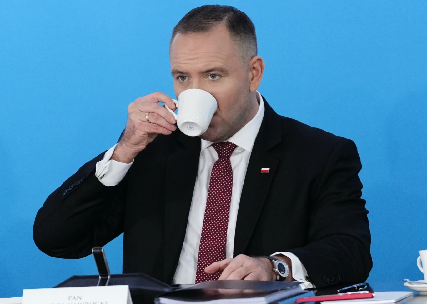 Mężczyzna w garniturze i czerwonym krawacie siedzi przy stole na tle niebieskiej ściany i pije z białej filiżanki. Na stole znajdują się dokumenty, mikrofon oraz filiżanka.