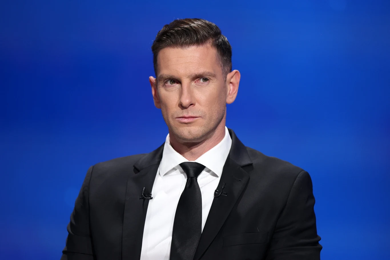 Łukasz Kadziewicz Elegancko ubrany mężczyzna w garniturze i czarnym krawacie na tle niebieskiego tła, z poważnym wyrazem twarzy i mikrofonami przypiętymi do marynarki.