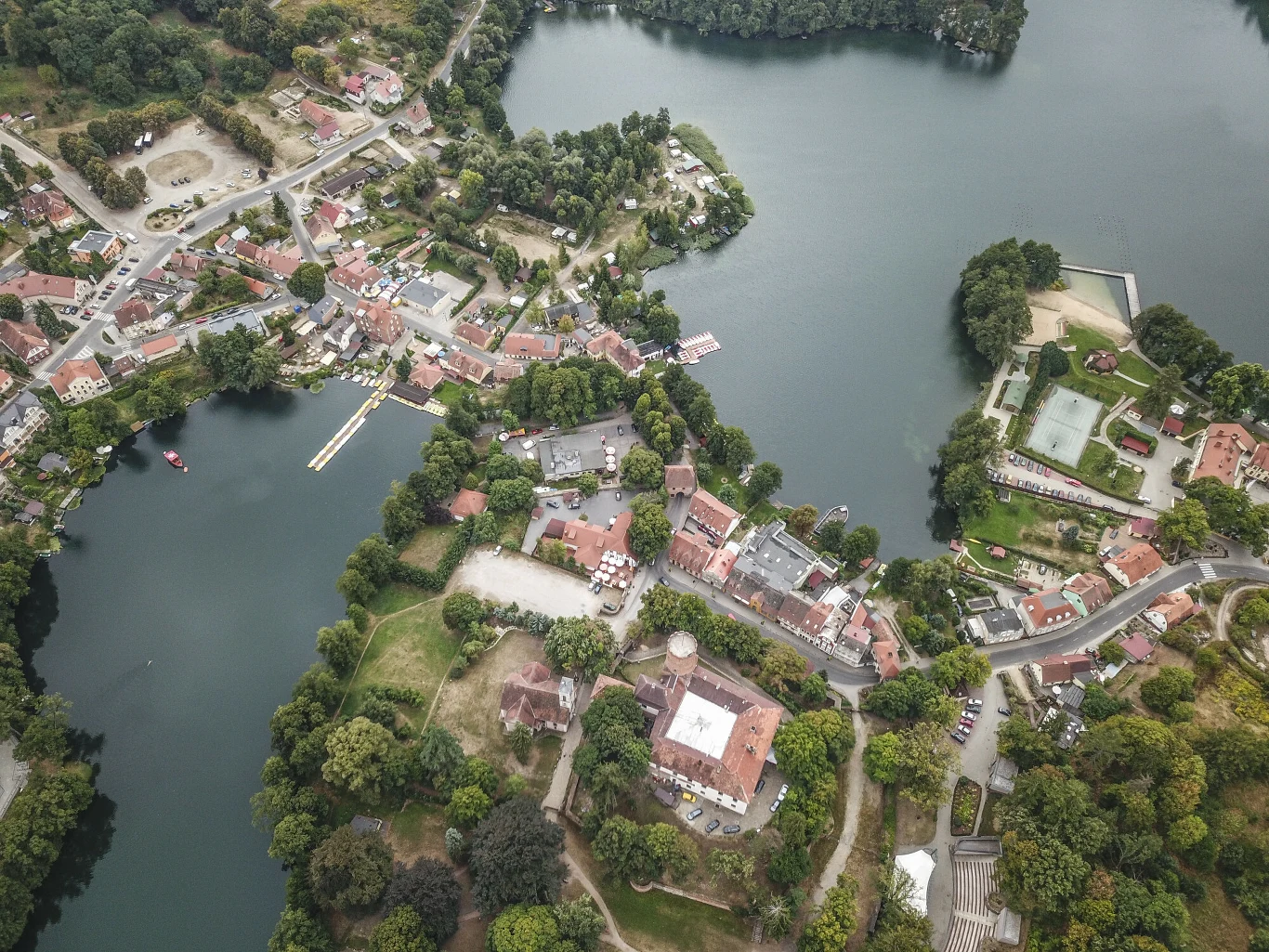 Panorama miasta z lotu ptaka z licznymi budynkami otoczonymi zielenią oraz dużym jeziorem, widać także drogi, most i przystań wodną.