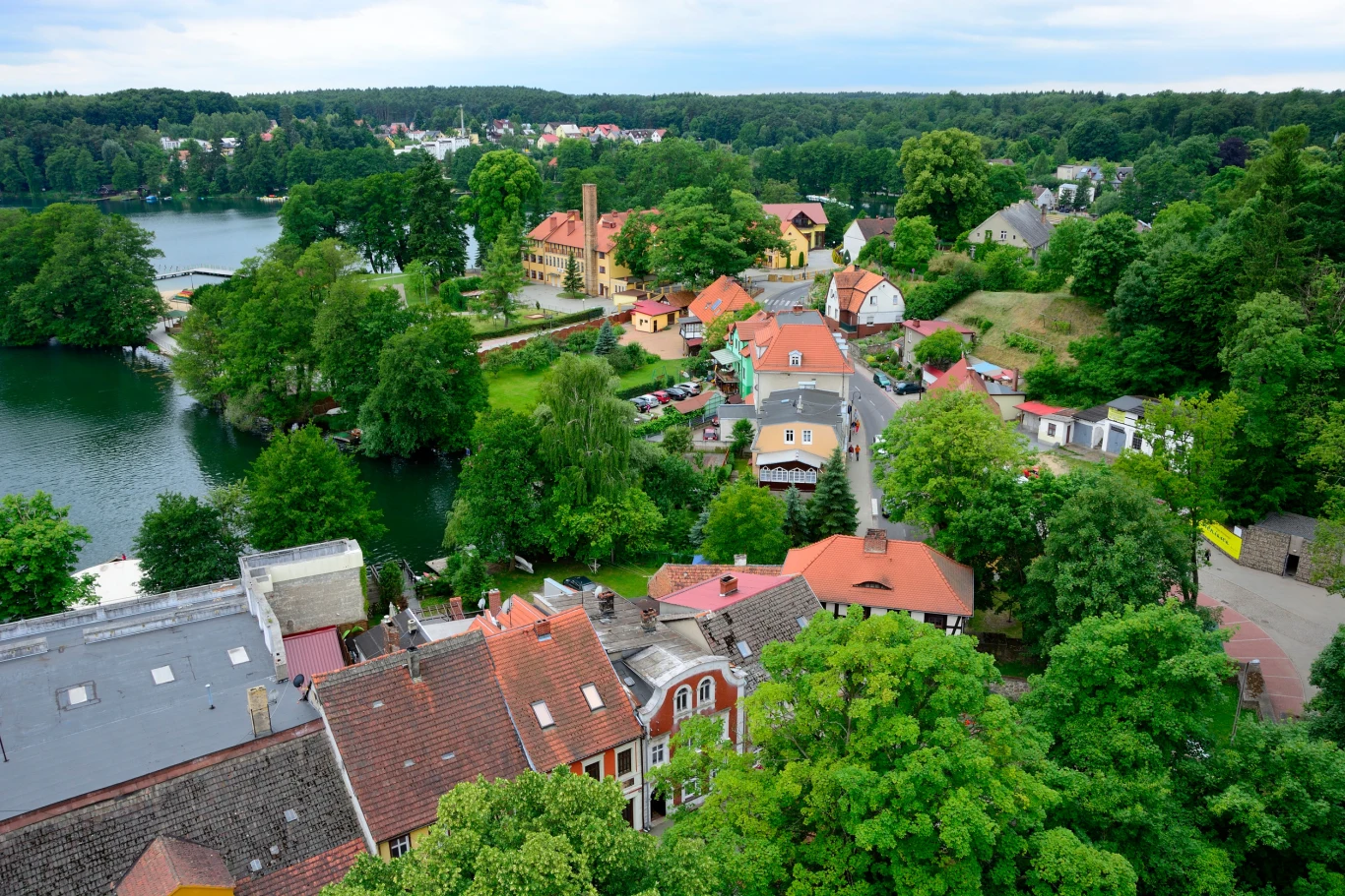 Kolorowe budynki w otoczeniu zieleni i jeziora, spokojne uliczki i zabytkowy pałac w centrum. Południowa część kadru wypełniona dachami domów, w tle rozległy las i błękitna woda jeziora.