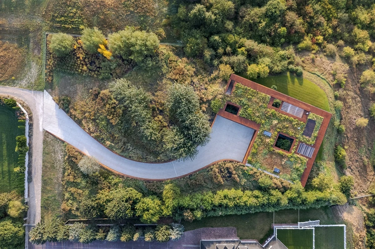 Budynek o nowoczesnej architekturze z zielonym dachem, otoczony gęstą roślinnością i drzewami. Do domu prowadzi zakręcona droga, a całość znajduje się w naturalnym, zadrzewionym terenie.