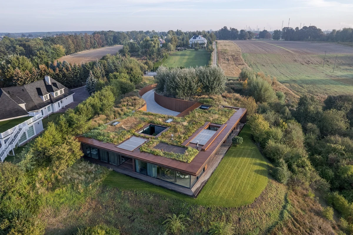 Nowoczesny budynek jednorodzinny z płaskim dachem pokrytym zielenią, otoczony bujną roślinnością i polami. W tle widoczne inne domy oraz linia drzew oddzielająca zabudowania od pól uprawnych.