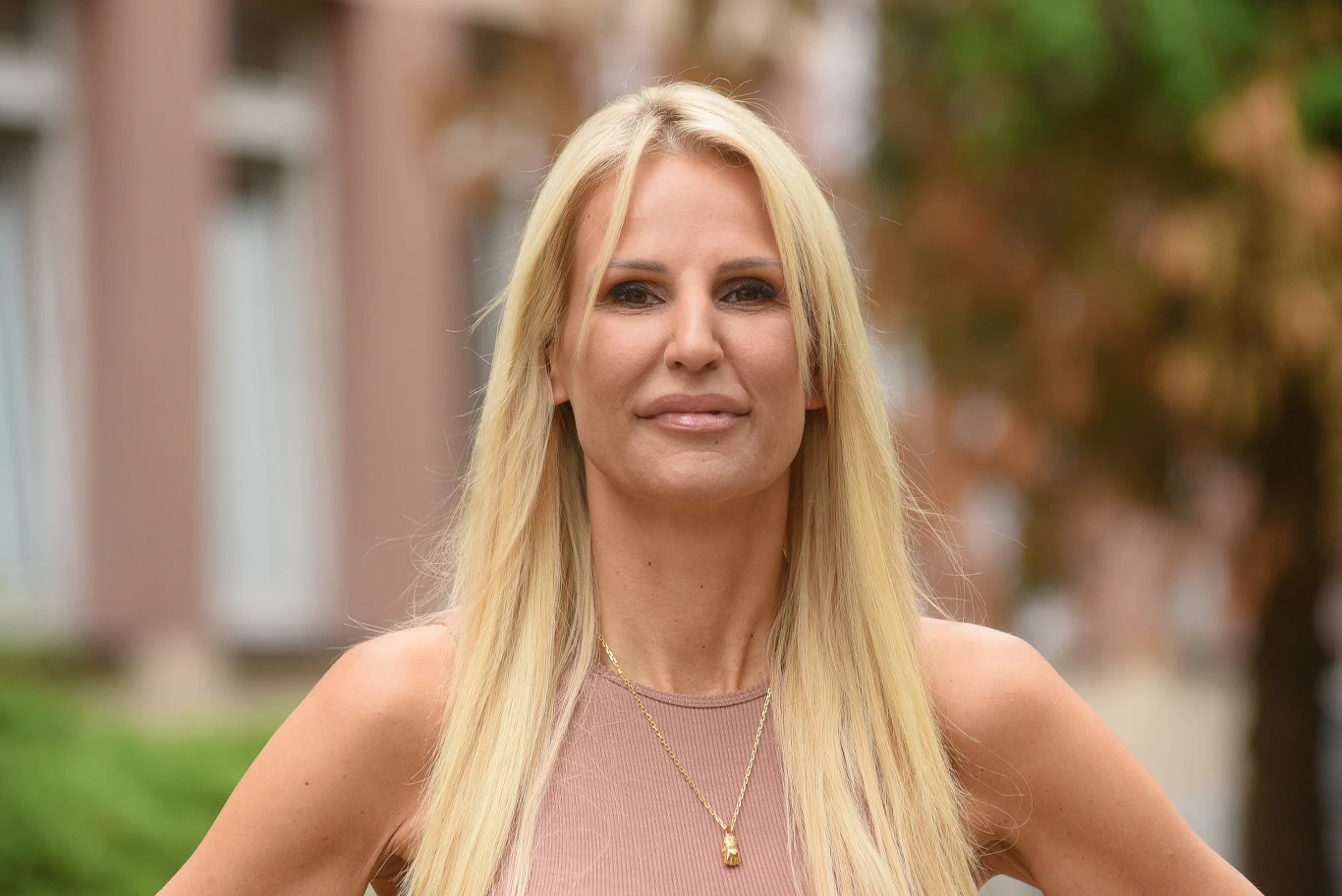 Dominika Tajner Blondynka o długich prostych włosach, ubrana w beżowy top, stoi przed budynkiem z rozmytym tłem roślinności oraz okien.