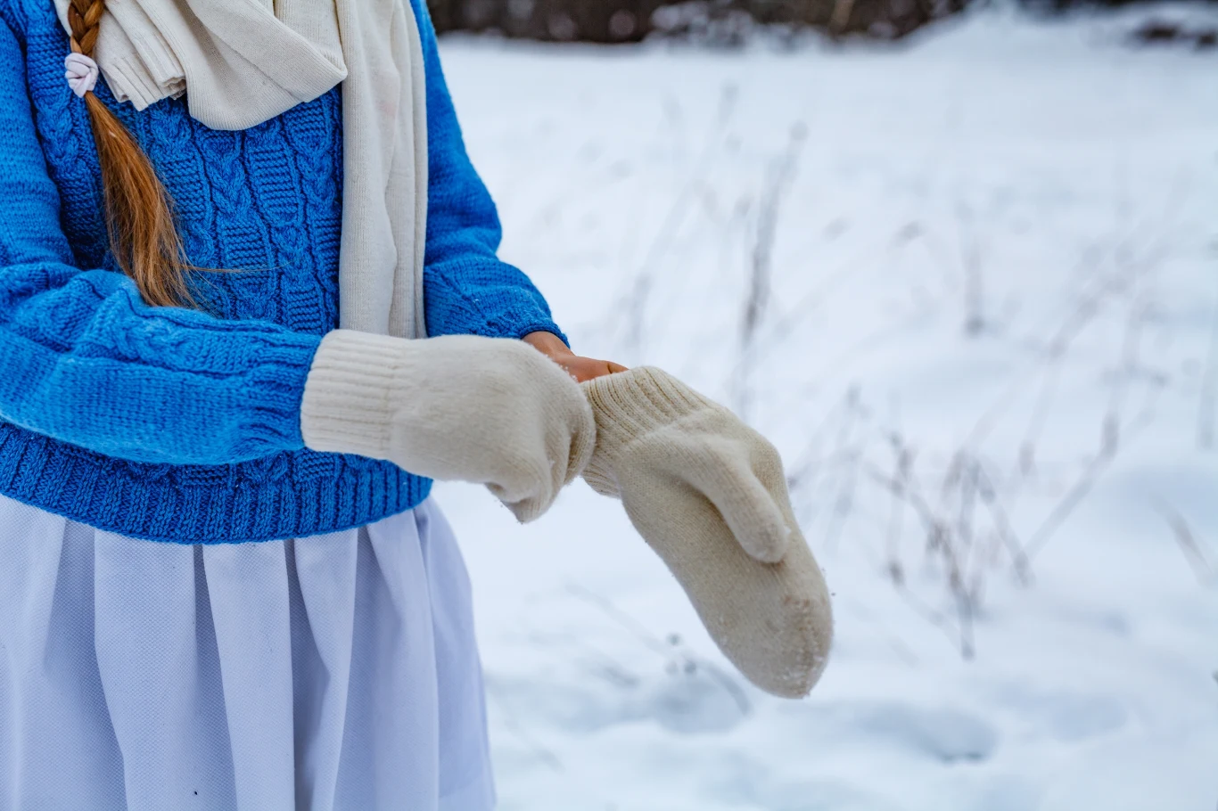 Zmarznięte dłonie od wilgoci i mrozu? Sprawdź ten trik Dziewczynka ubrana w niebieski sweter, białą spódnicę oraz ciepły szalik zakłada jasne wełniane rękawiczki. W tle rozciąga się zimowy, ośnieżony krajobraz.