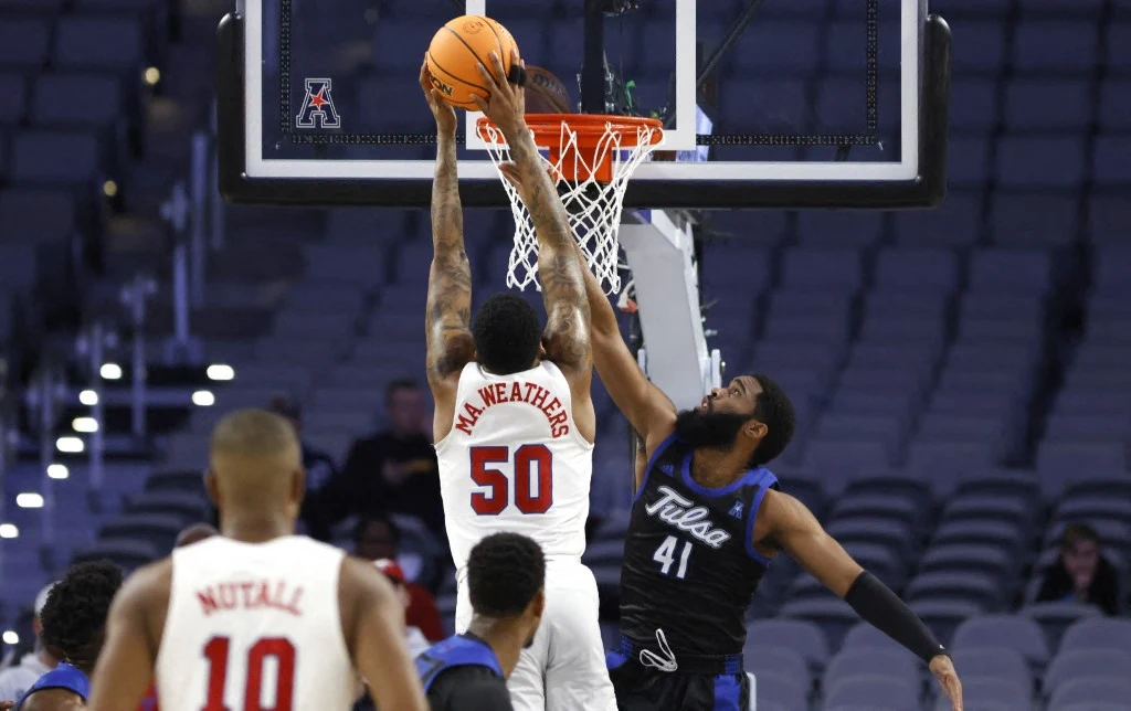 Zawodnik w białej koszulce z numerem 50 wykonuje wsad do kosza podczas meczu koszykówki, próbując zdobyć punkty pomimo obrony gracza w czarnej koszulce z numerem 41. W tle widoczni są inni zawodnicy oraz puste trybuny.