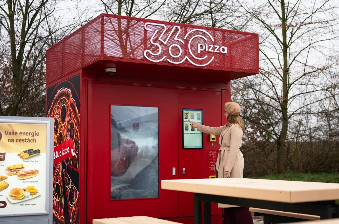 Automat do sprzedaży pizzy w czerwonym kolorze z wyświetlaczem dotykowym obsługiwany przez elegancko ubraną kobietę, obok znajduje się menu z ofertą potraw. W tle widoczne nagie drzewa.