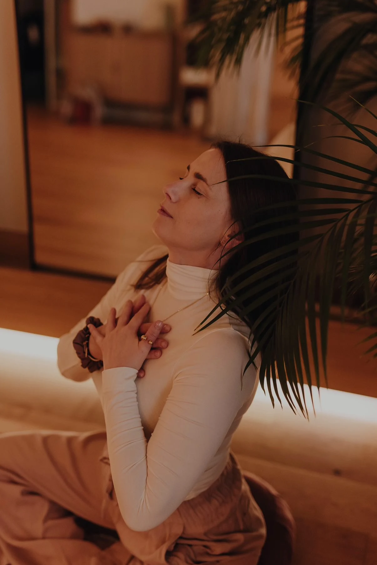 Kobieta w jasnym swetrze siedzi ze skrzyżowanymi nogami w pozycji relaksacyjnej, trzyma dłonie skrzyżowane na klatce piersiowej, ma zamknięte oczy, otacza ją spokojna atmosfera wnętrza z ciepłym oświetleniem i rośliną na pierwszym planie.