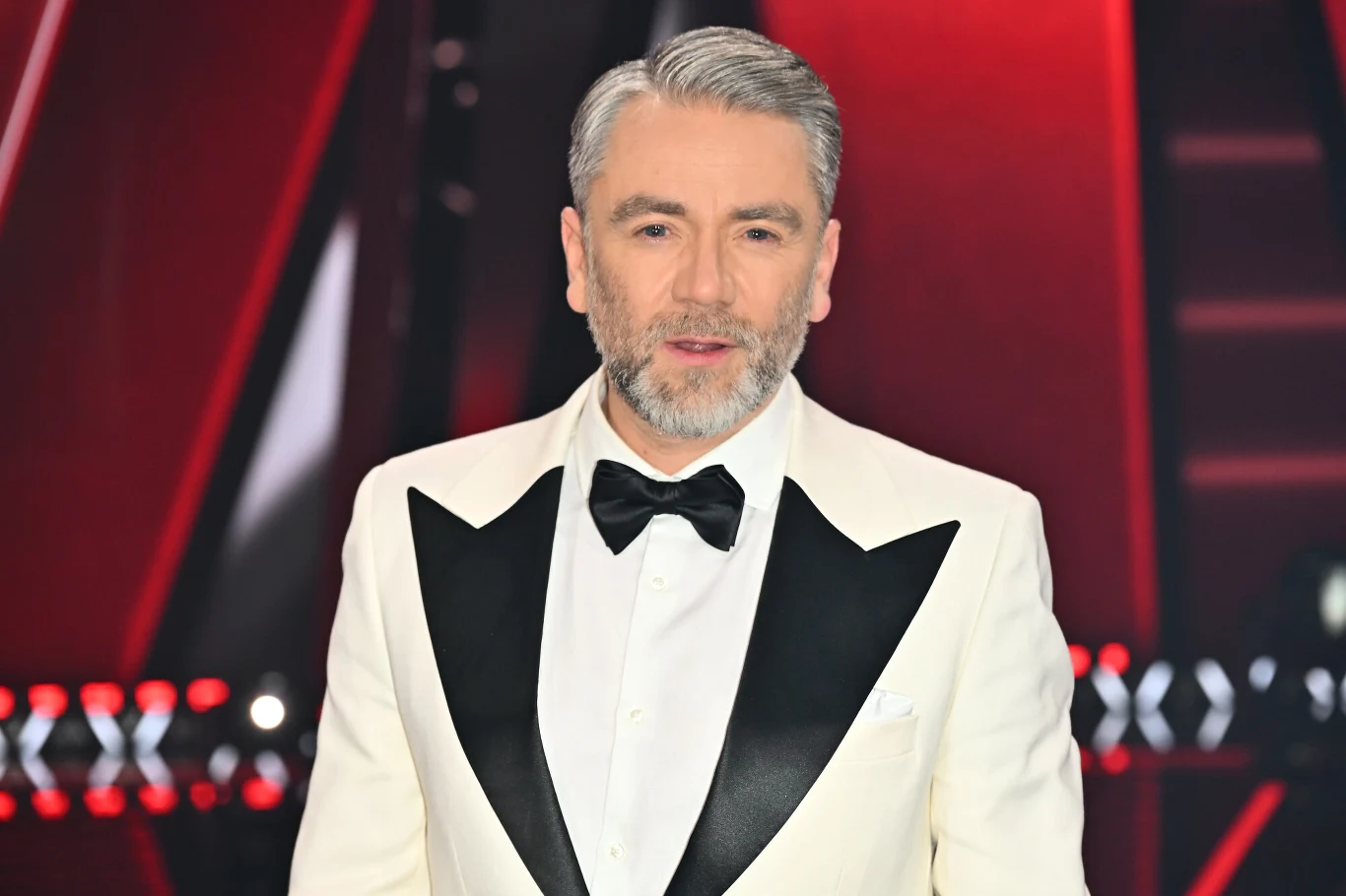 Kuba Badach promuje swój czwarty solowy album "Radio Edit" Mężczyzna z siwymi włosami i brodą w eleganckim białym smokingu z czarnymi klapami i muchą, stojący na tle kontrastujących czerwono-czarnych elementów scenicznych.