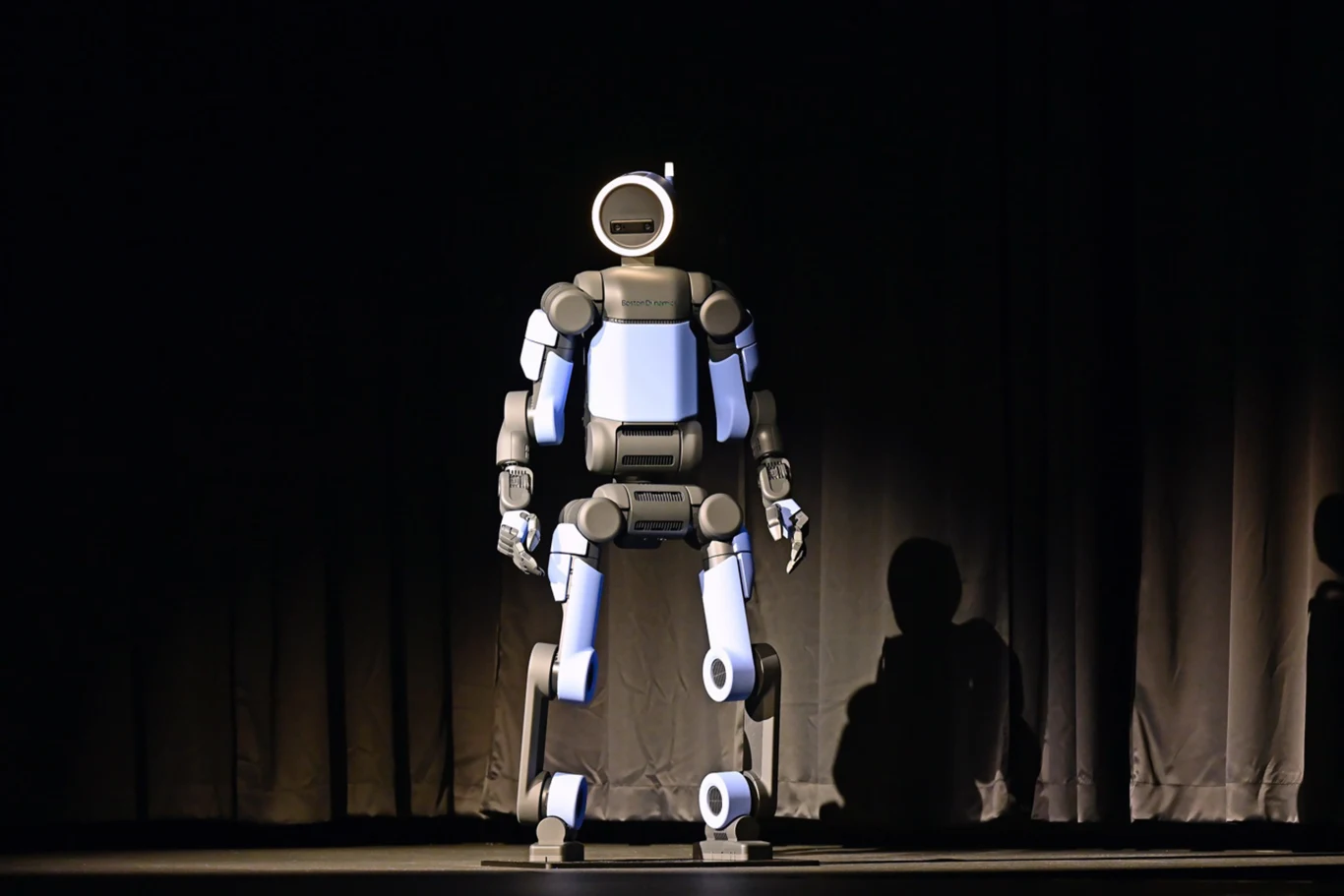 Duży humanoidalny robot o niebiesko-czarnych panelach stoi na scenie, oświetlony światłem, na tle ciemnych kotar. Obok widać cień postaci sugerujący obecność człowieka przy robocie.