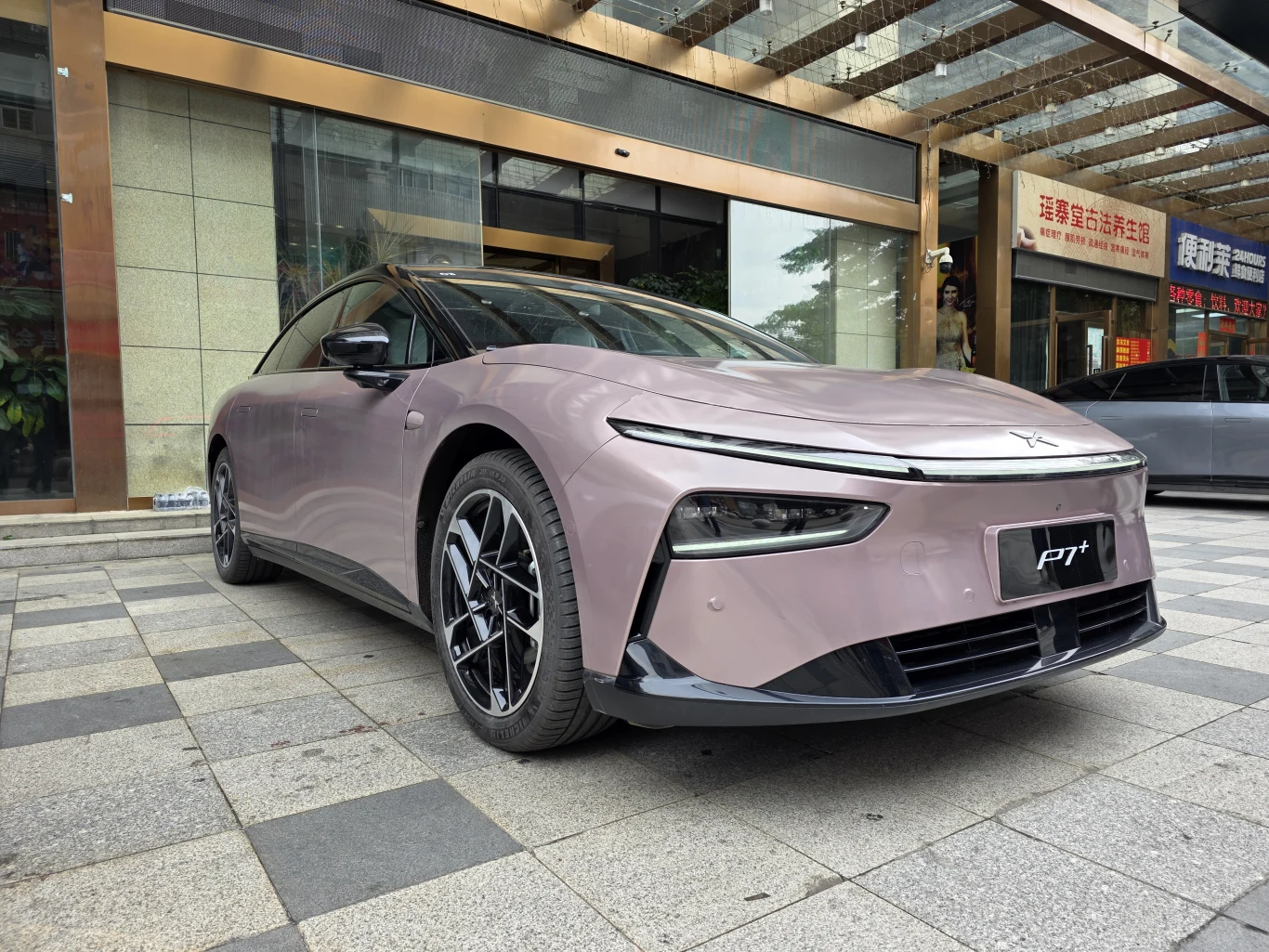 Nowoczesny samochód elektryczny w jasnofioletowym kolorze zaparkowany na chodniku przed przeszklonym budynkiem z widocznymi szyldami w języku chińskim. Auto cechuje się aerodynamiczną sylwetką, eleganckimi felgami i wąskimi reflektorami LED.