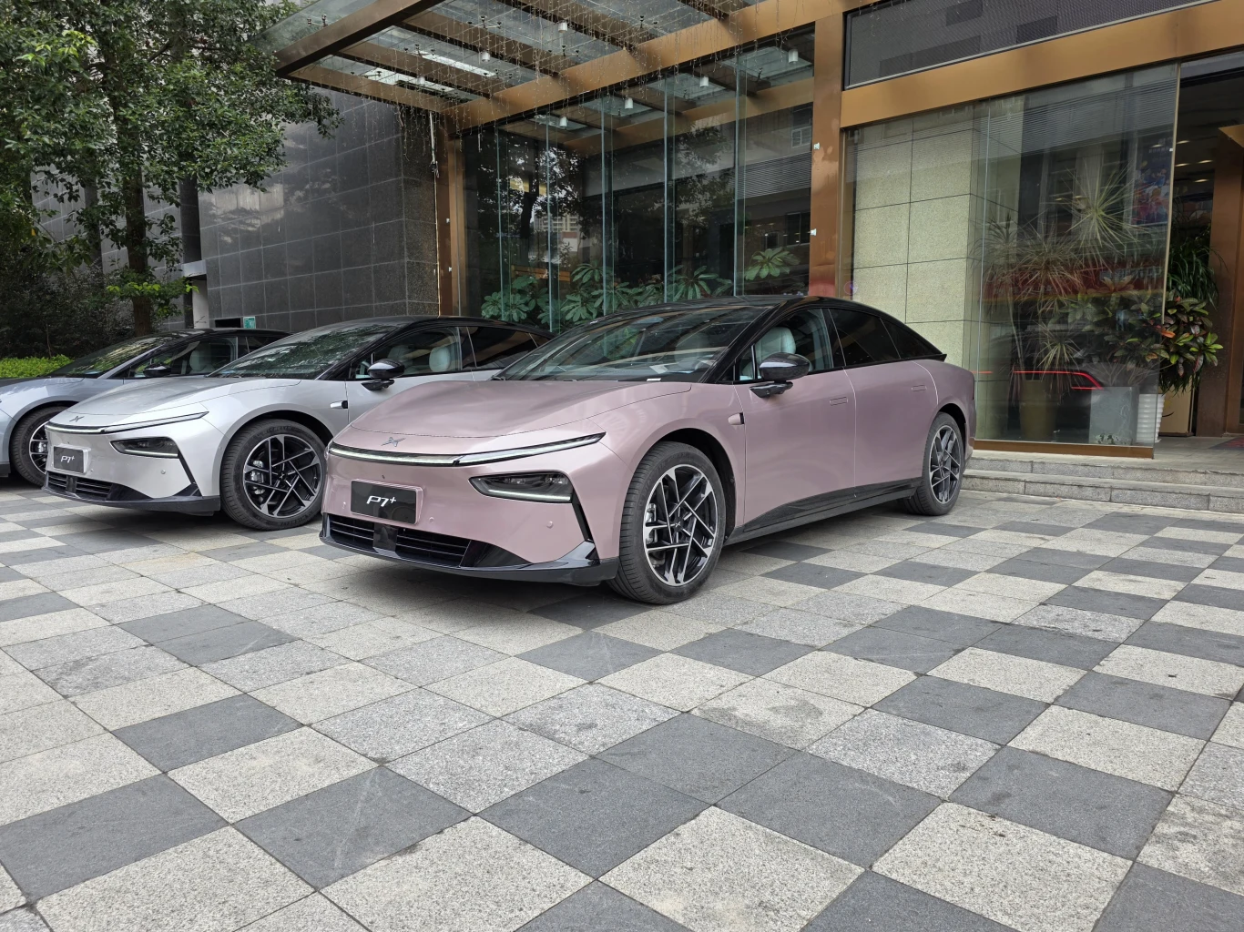 Dwa nowoczesne samochody elektryczne zaparkowane przed przeszklonym budynkiem, oba pojazdy wyróżniają się aerodynamiczną sylwetką i minimalistycznym designem, otoczenie stanowi elegancka, wyłożona płytkami przestrzeń.