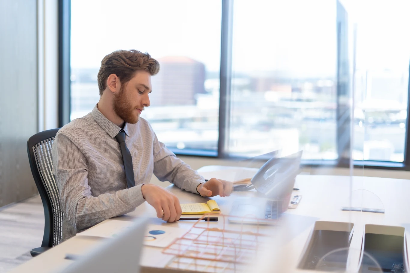 Mężczyzna w koszuli i krawacie siedzi przy biurku w nowoczesnym biurze, skupiając się na przeglądaniu dokumentów, przez okna widoczny rozmyty krajobraz miasta.