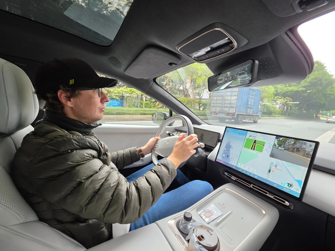 Mężczyzna w czapce z daszkiem prowadzi nowoczesny samochód wyposażony w duży ekran dotykowy z nawigacją, na konsoli centralnej ustawiony napój, za szybą widoczna droga i ciężarówka.