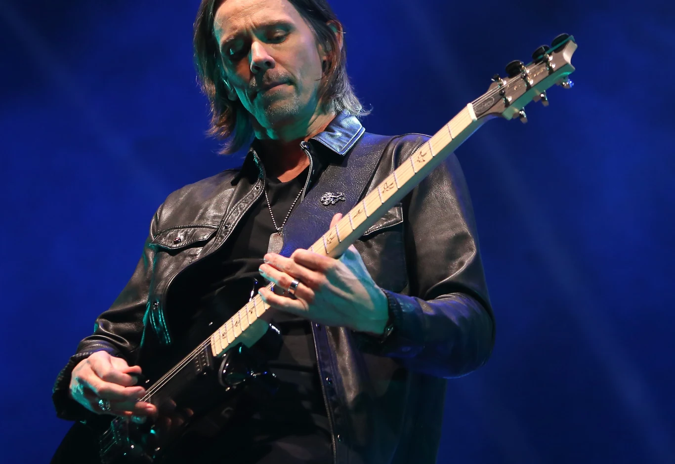 Alter Bridge wystąpi w Gliwicach 27 stycznia Muzyk grający na gitarze elektrycznej ubrany w czarną skórzaną kurtkę, skoncentrowany na swoim występie na tle niebieskiego oświetlenia scenicznego.