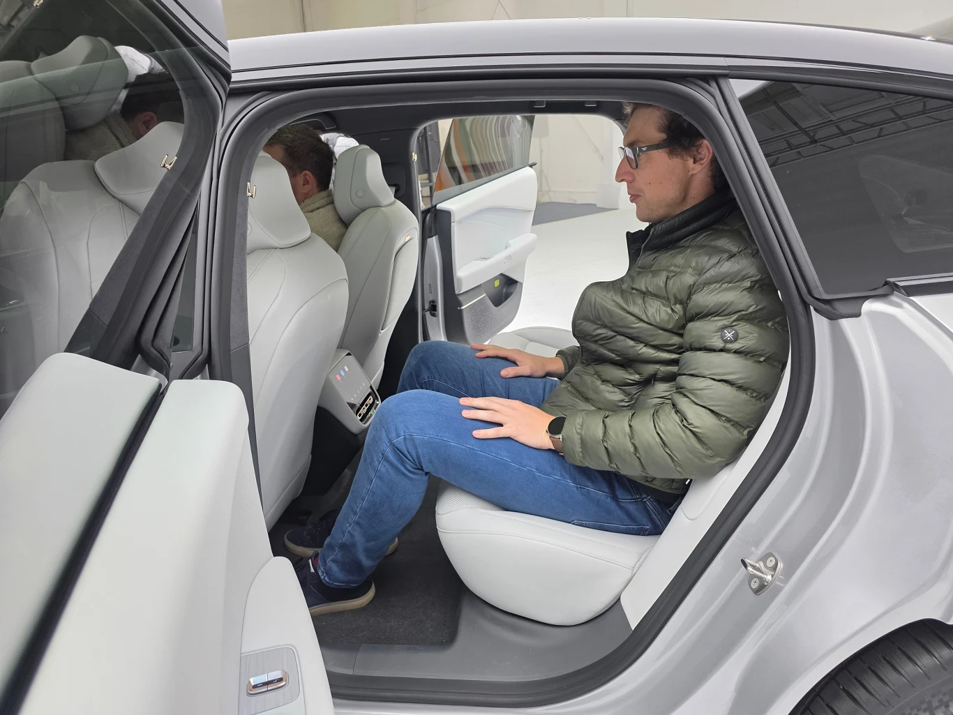Mężczyzna siedzący na tylnym siedzeniu nowoczesnego samochodu, ubranego w zieloną kurtkę i niebieskie jeansy, testuje przestrzeń na nogi oraz wygodę siedzenia, z wyraźnie uchylonymi drzwiami i jasnym wnętrzem pojazdu.