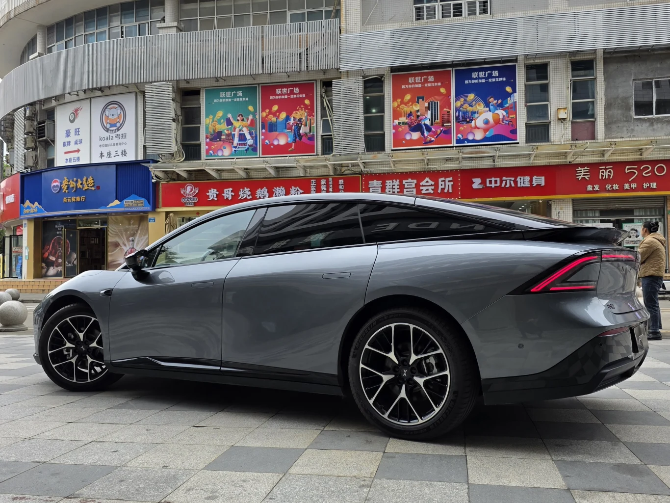 Nowoczesny szary samochód osobowy o sportowej sylwetce zaparkowany na tle miejskich budynków z kolorowymi reklamami i witrynami sklepów.