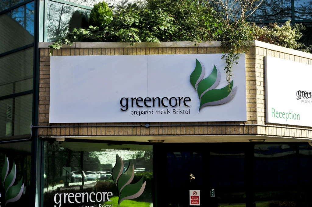 Oddział firmy Greencore produkującej gotowe kanapki Fasada budynku z szyldem firmy Greencore oraz napisem 'prepared meals Bristol', ozdobiona roślinnością, z fragmentem wejścia i odbiciem zieleni w szybie.