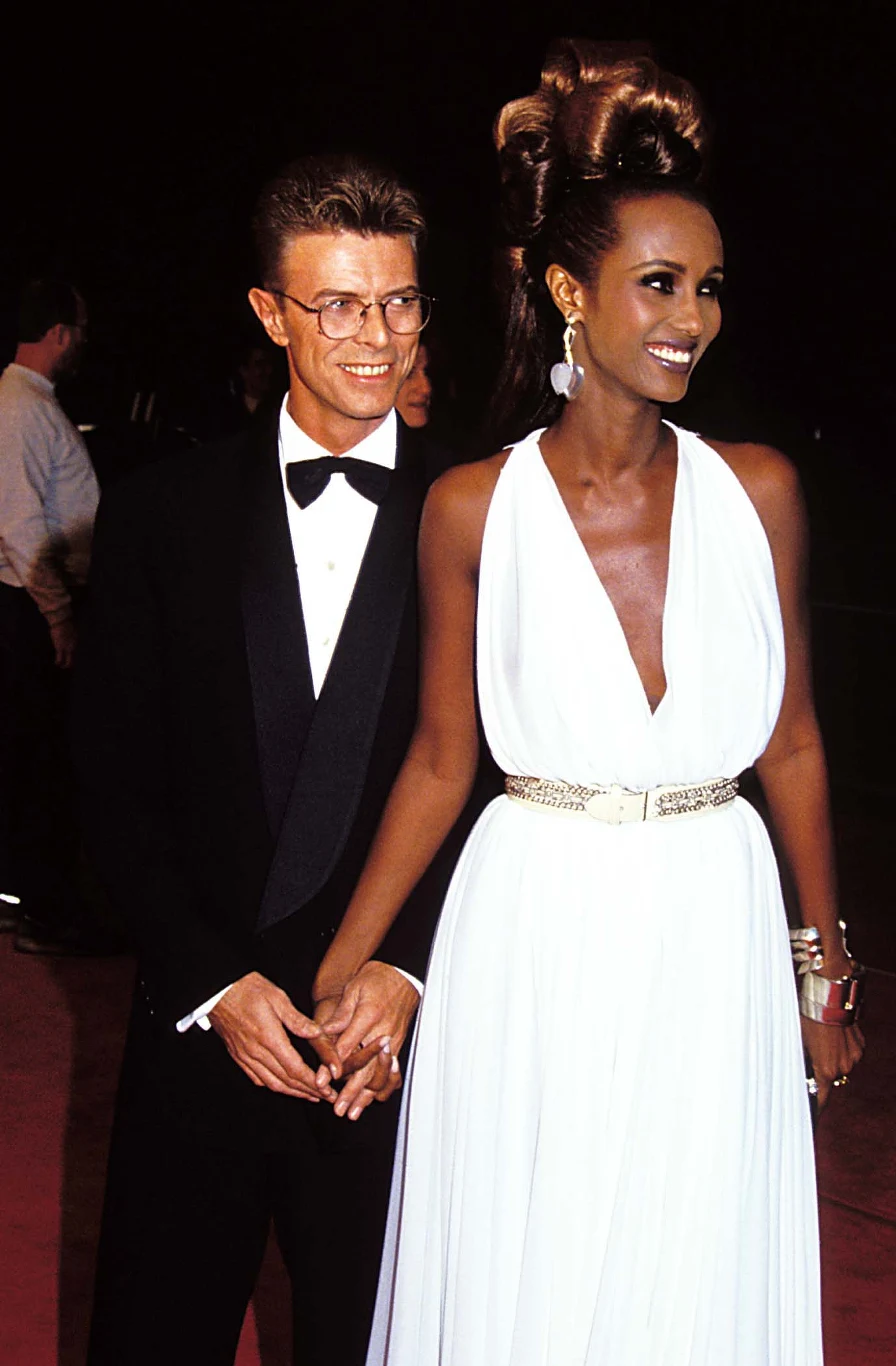 Elegancka para trzymająca się za ręce, mężczyzna w smokingu i okulary, kobieta w długiej białej sukni z efektowną fryzurą i biżuterią. Oboje uśmiechnięci, stoją na tle czerwonego dywanu w otoczeniu innych osób.