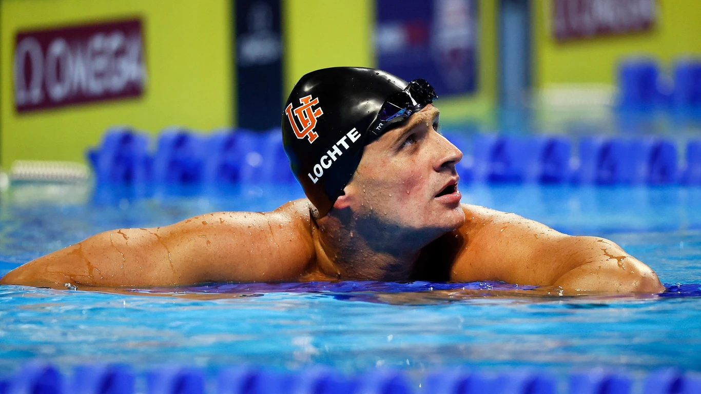 Ryan Lochte Pływak w czepku i okularach pływackich odpoczywa przy krawędzi basenu, jego ciało unosi się na wodzie, a wzrok skierowany jest w górę. W tle widoczne są elementy wyposażenia basenu sportowego.