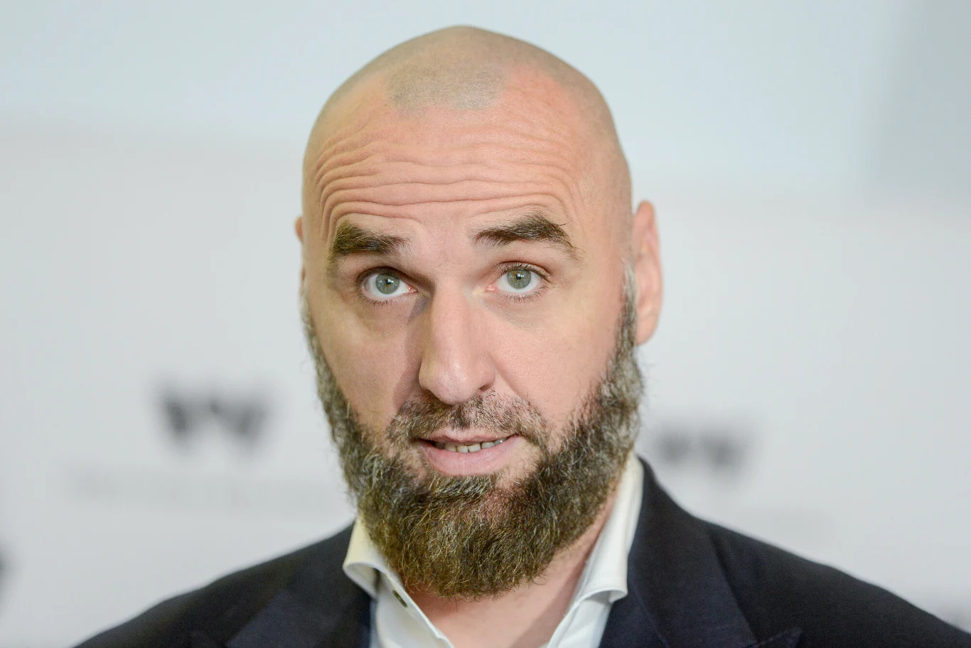 Mężczyzna z łysą głową i gęstą brodą w garniturze, patrzący prosto w obiektyw, jasne tło z rozmytymi elementami graficznymi