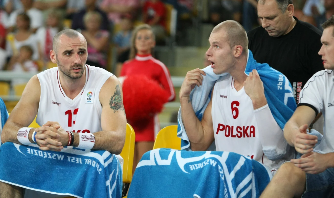 Koszykarze polskiej reprezentacji siedzący na ławce rezerwowych z ręcznikami na ramionach, w tle widoczna cheerleaderka oraz kibice na trybunach.